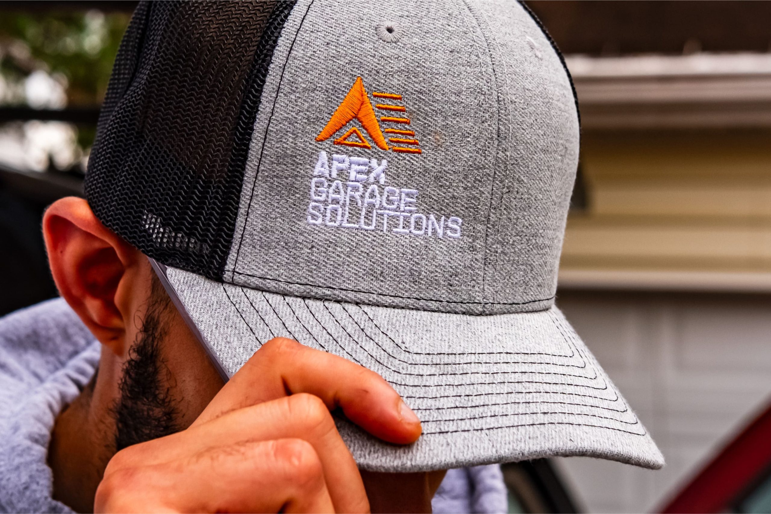 Close-up of a grey trucker cap with black mesh back, featuring the embroidered orange and white APEX Garage Solutions chevron logo.