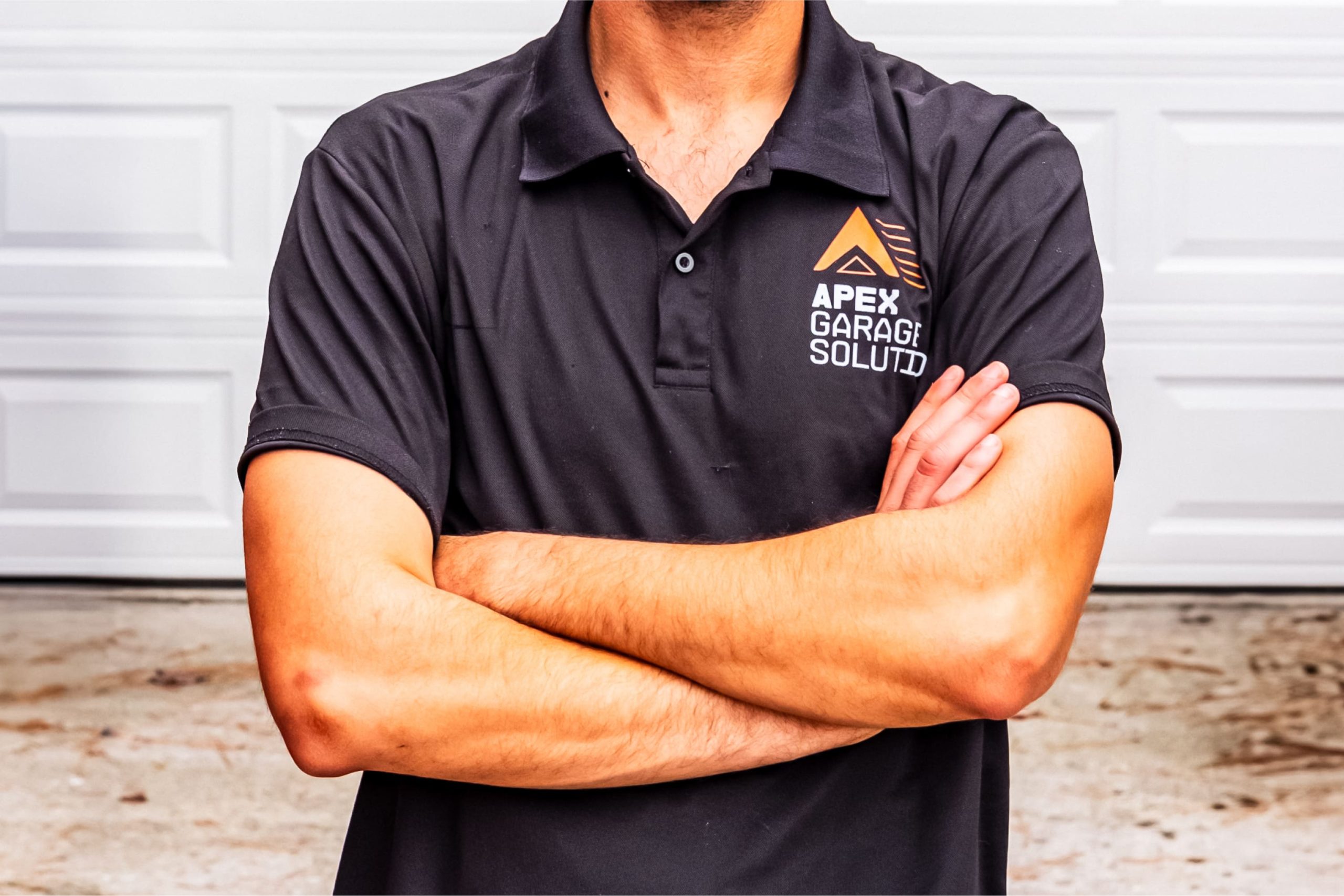 Technician standing in front of a white residential garage door wearing a black polo shirt with the APEX Garage Solutions logo on the left chest.