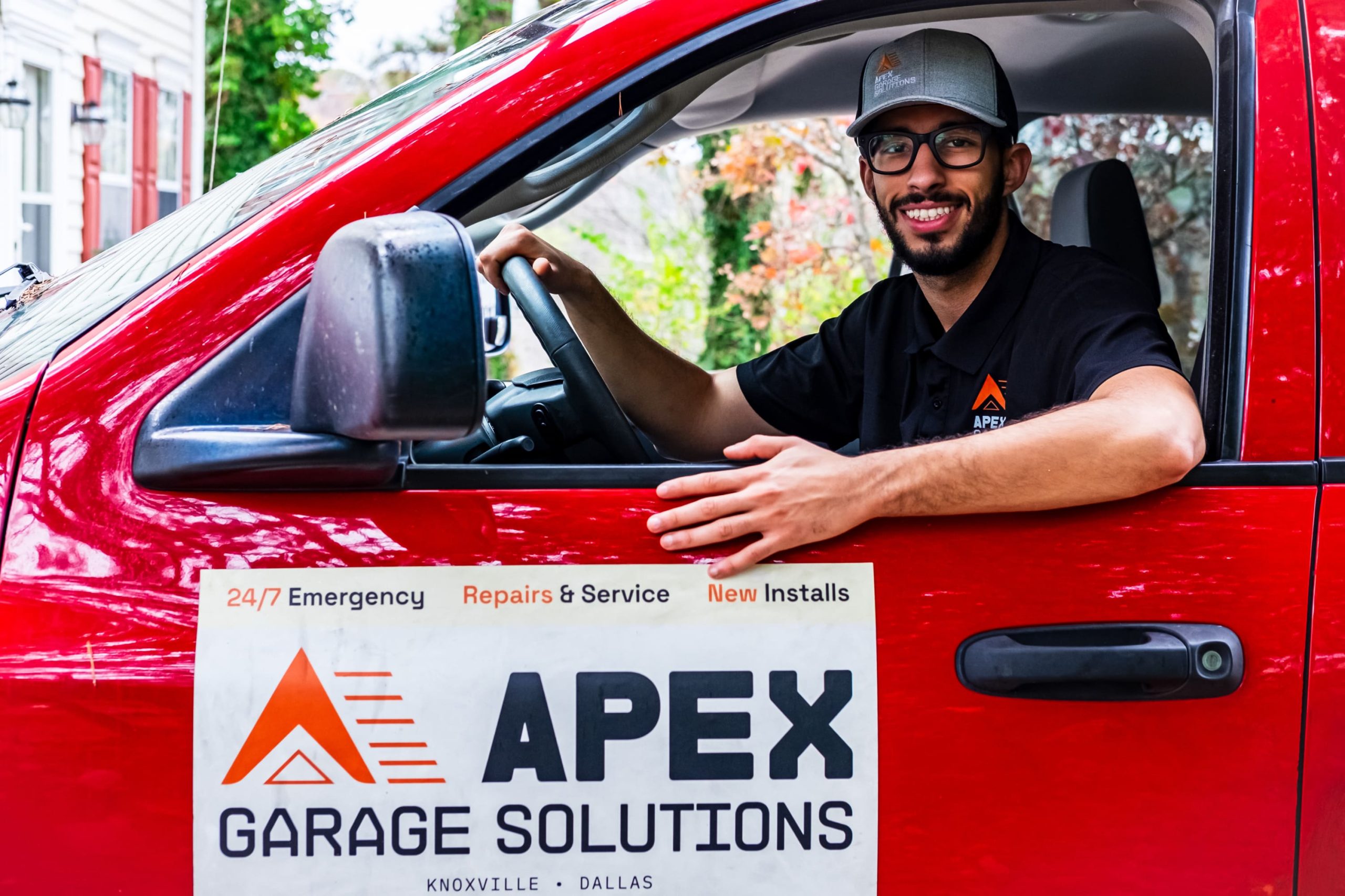 Technician seated in the driver's seat of a red work vehicle with an APEX Garage Solutions door magnet reading 24/7 Emergency Repairs & Service and listing Knoxville and Dallas locations.