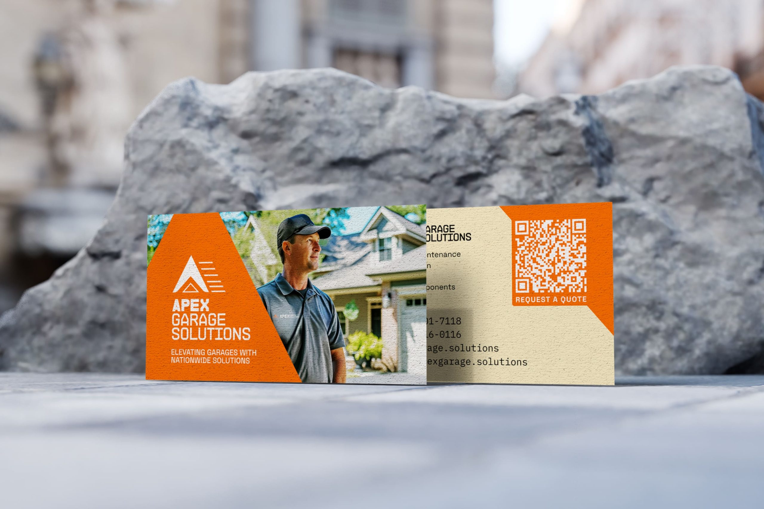 Business cards standing on a stone surface showing the front card with technician portrait and orange banner, and the back card with service lists, phone numbers, and a QR code.