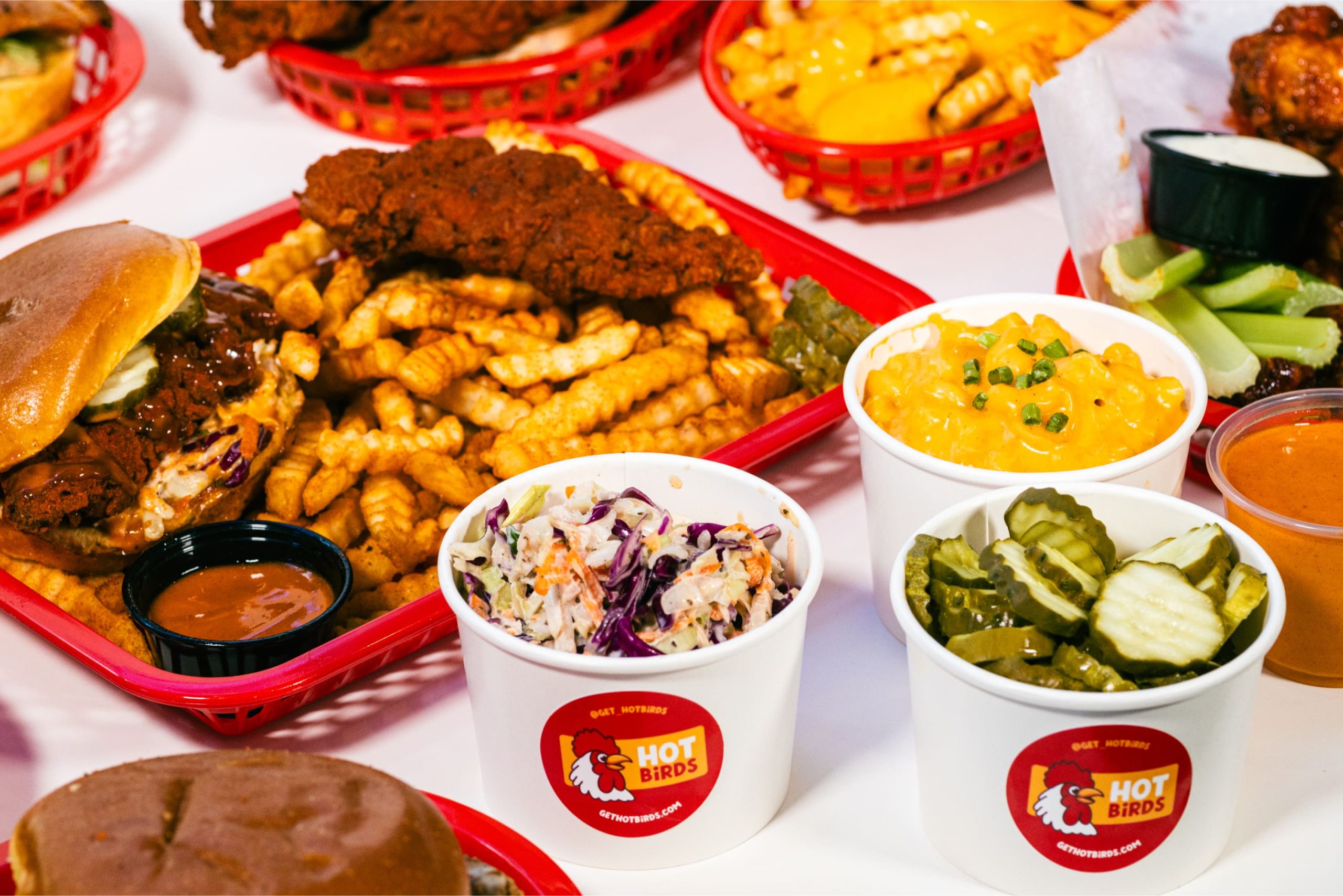 Overhead flat-lay view of a hot chicken sandwich, fried tenders in a red basket with crinkle fries, dipping sauce cups, mac and cheese, pickles, coleslaw, and celery.