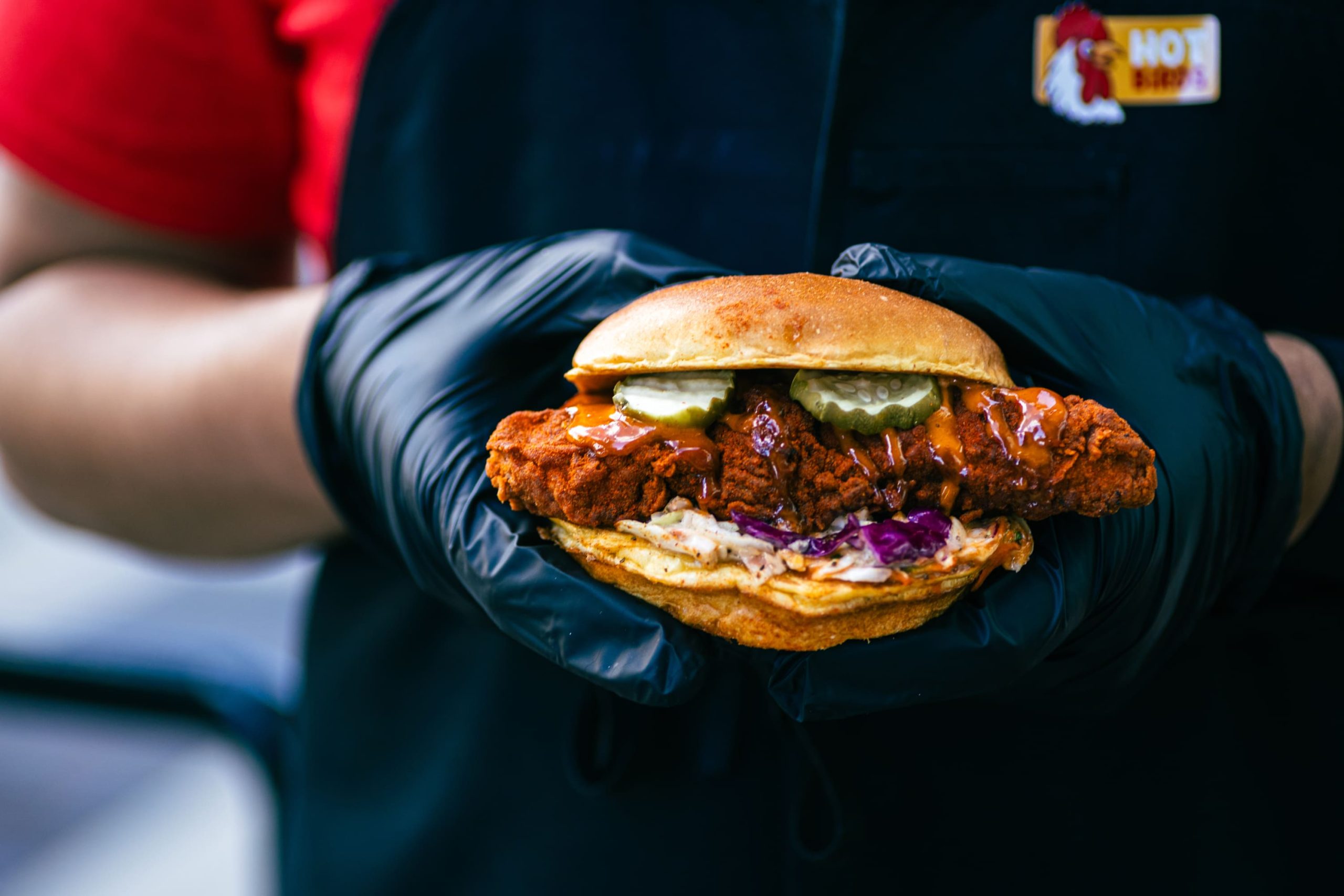 A hot chicken sandwich with fried chicken breast, pickles, and coleslaw on a toasted bun held in black-gloved hands by a team member in a branded black apron.