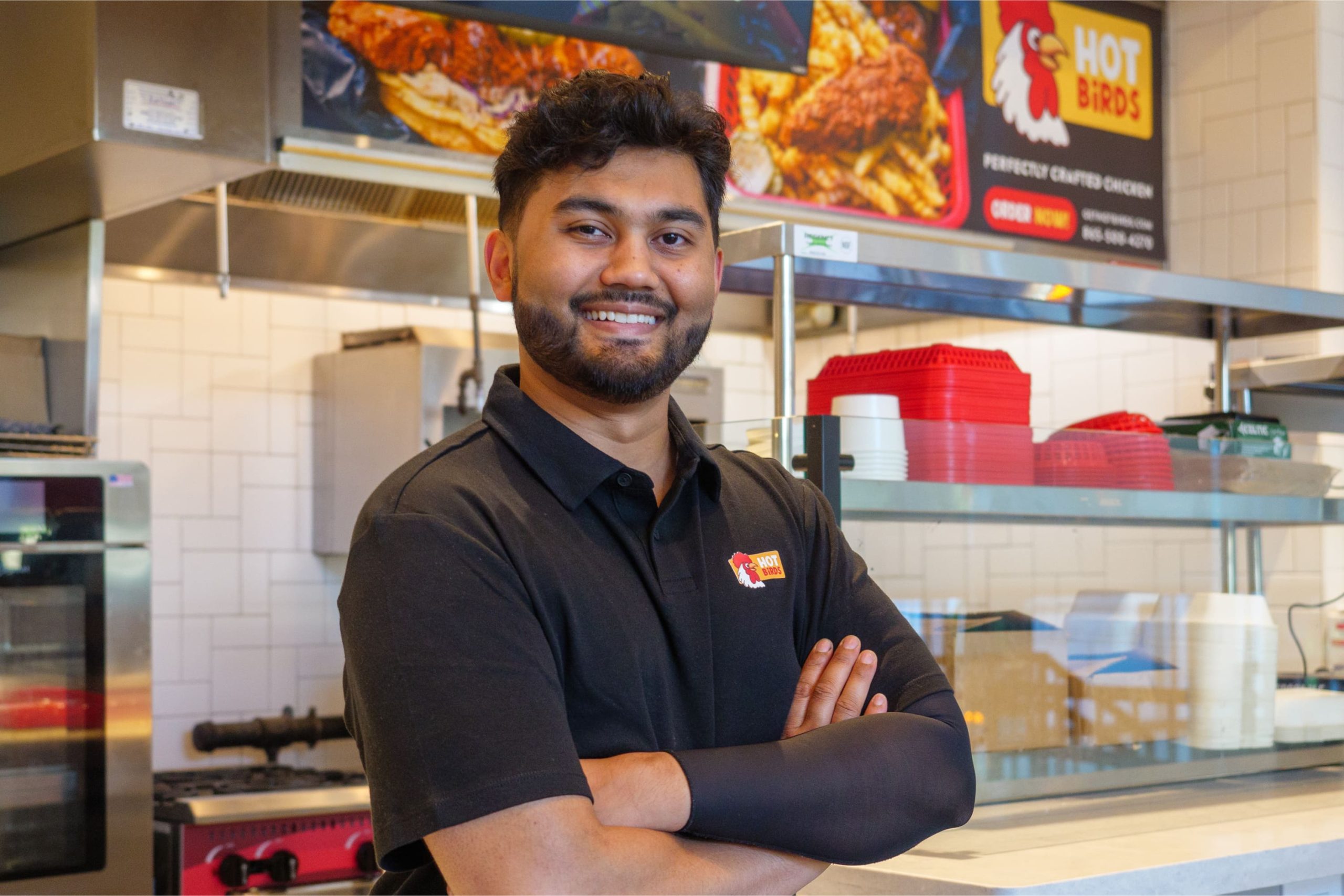 Team member in a black Hot Birds polo shirt standing with arms crossed in front of the kitchen service counter and digital menu displays.