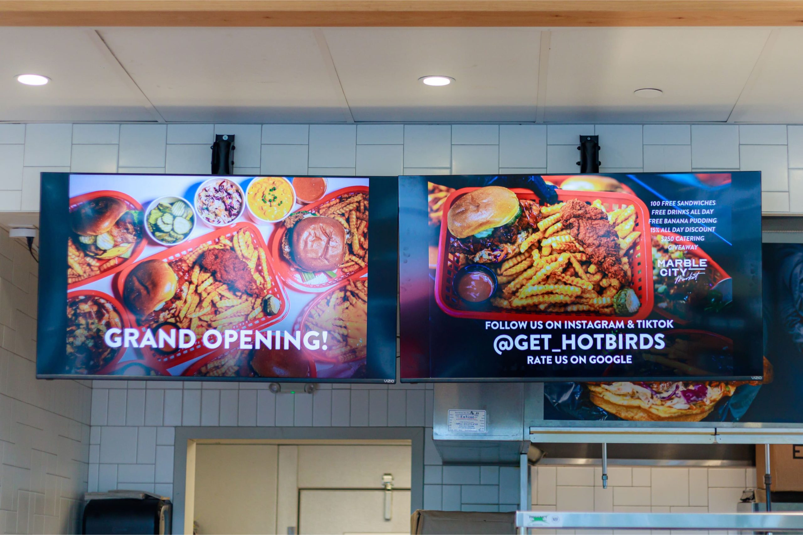 Two overhead digital screens displaying Grand Opening announcements, menu promotions, social media handle @get_hotbirds, and a Google review prompt.