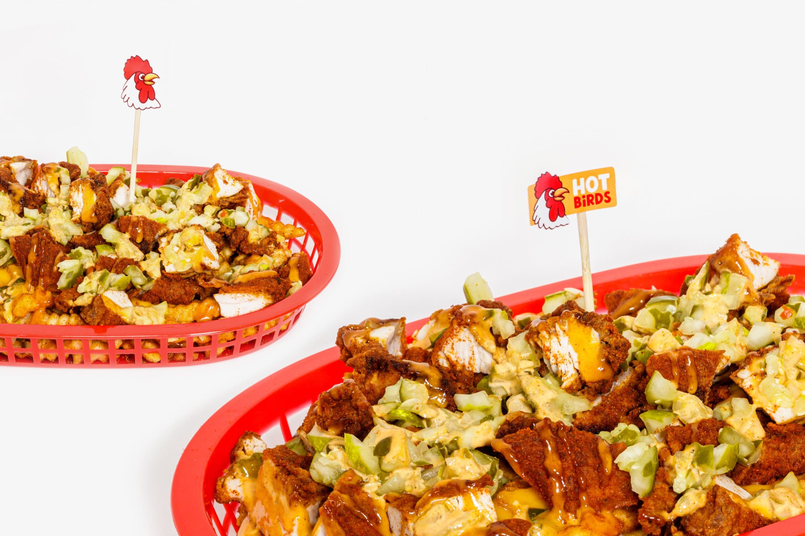 Red serving baskets filled with loaded crinkle fries and fried chicken bites topped with sauce, each garnished with a miniature Hot Birds flag on a wooden toothpick.
