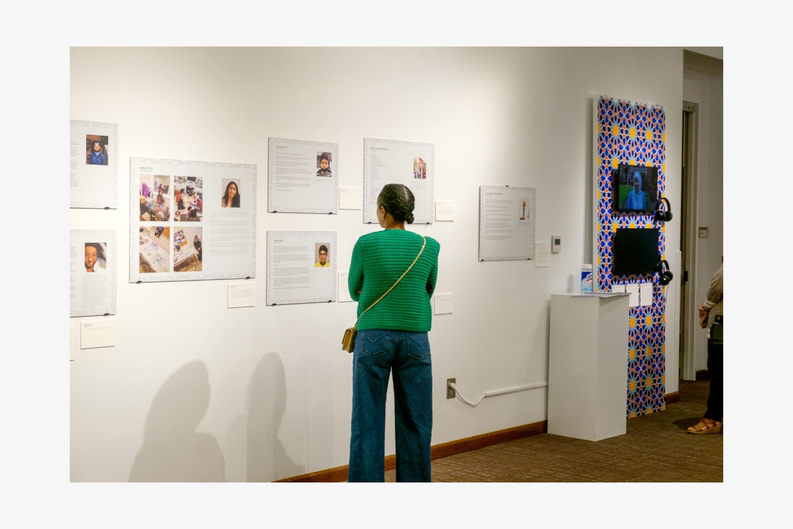 Exhibition visitor reading biographical wall panels next to a decorative panel mounted with two video screens and headphones.