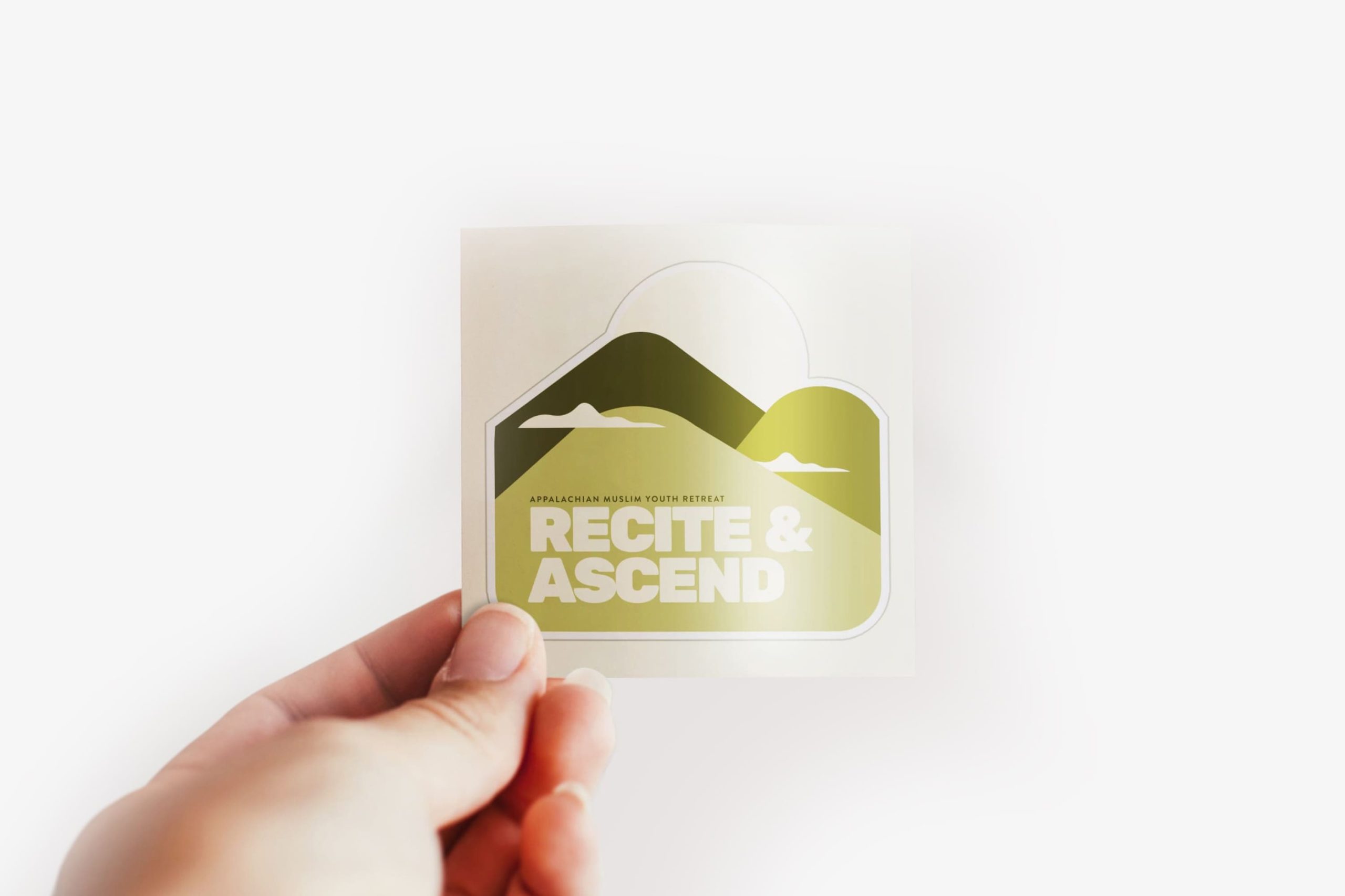 Hand holding a die-cut vinyl sticker of the olive green mountain peak and sun emblem with typography reading Appalachian Muslim Youth Retreat / Recite & Ascend.